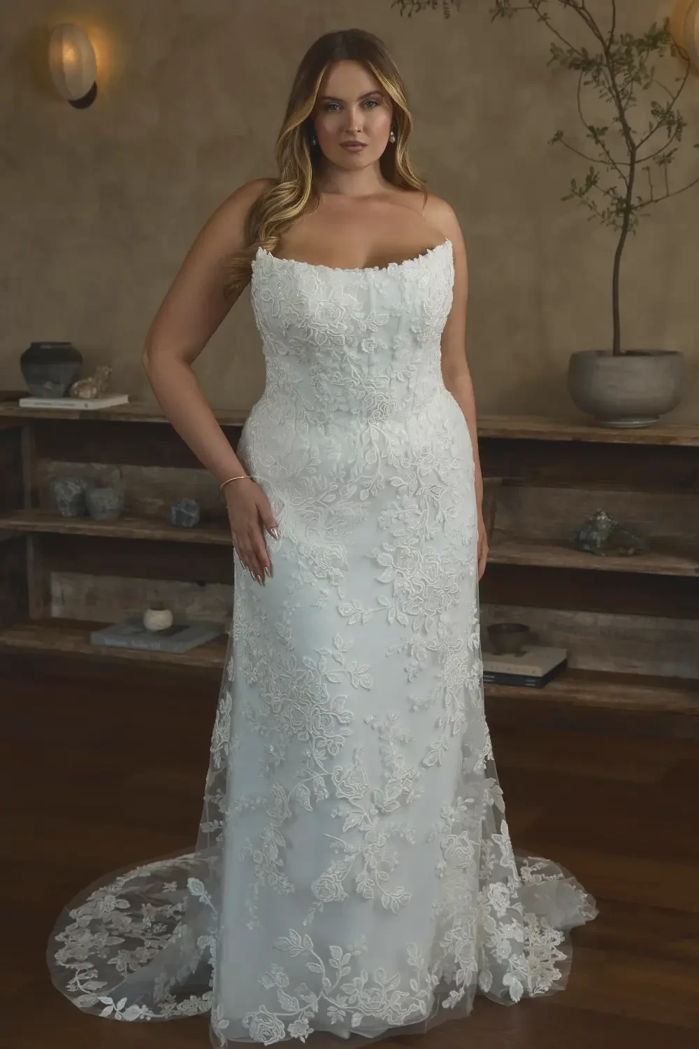 A woman in a fitted white wedding dress with floral lace detailing, standing elegantly in a room with a rustic atmosphere.