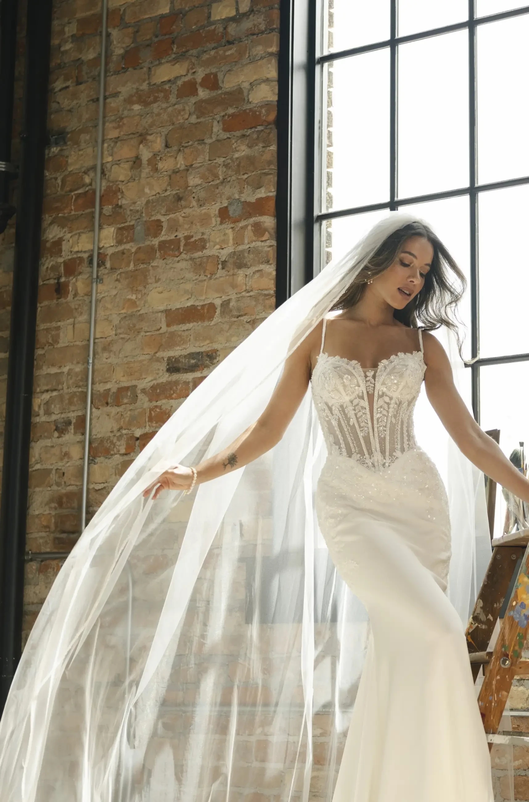 Bride in a flowing white wedding gown with intricate lace detail stands by a large window. Her long veil drapes elegantly, contrasting with a rustic brick wall.