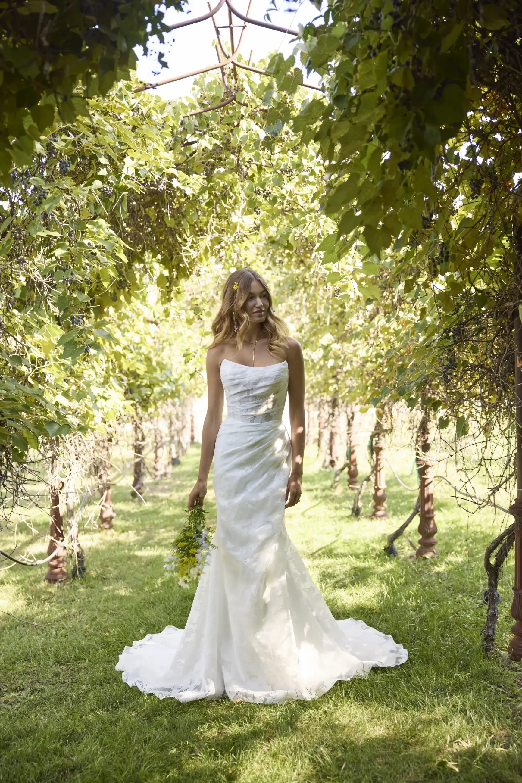 A bride in a white wedding dress stands in a vine-covered outdoor setting, surrounded by greenery. She holds a bouquet of flowers and looks serene.