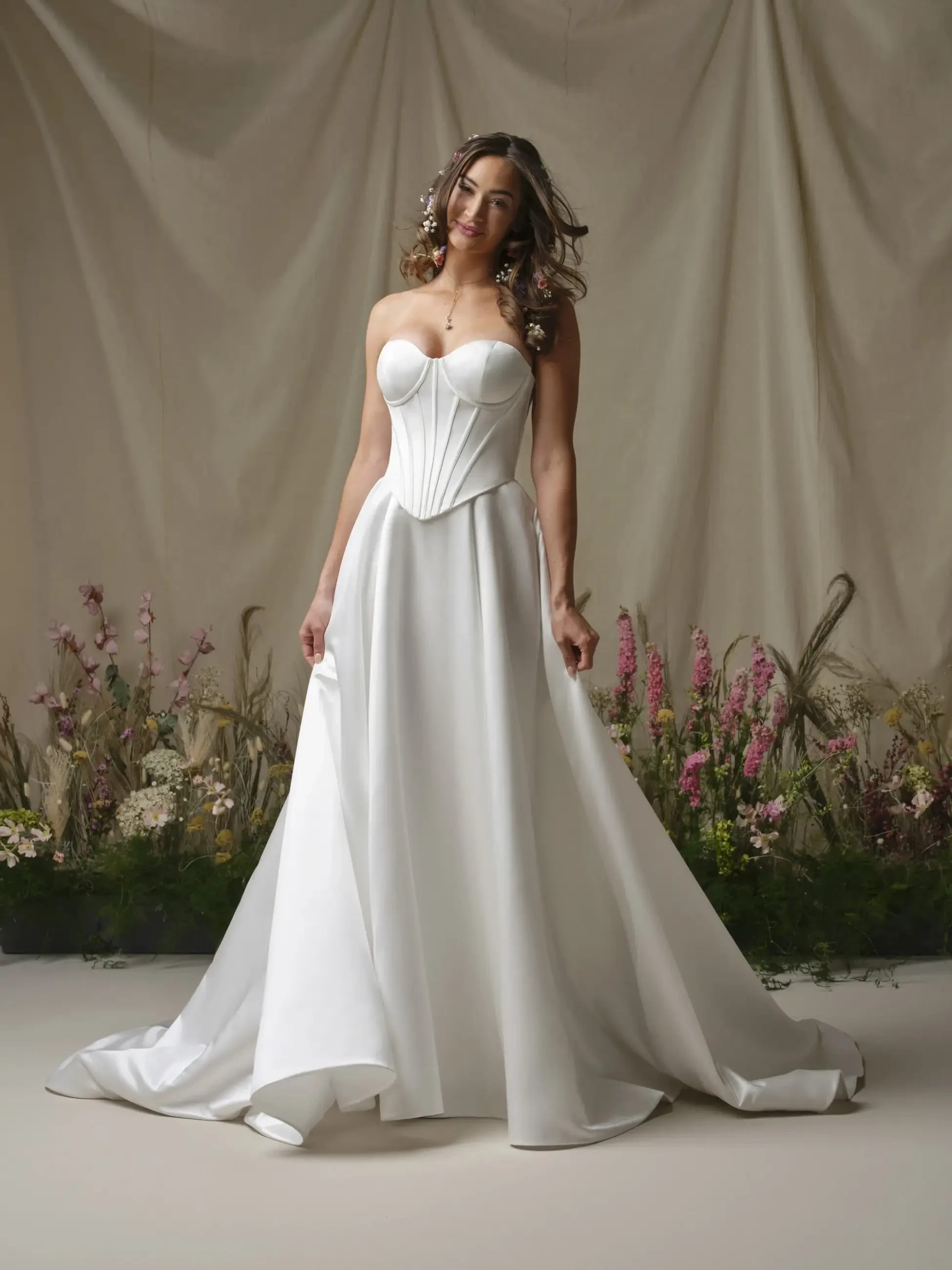 A woman in an elegant white strapless gown stands gracefully, surrounded by pink and white flowers. The backdrop is a soft, draped fabric, exuding a serene and romantic tone.