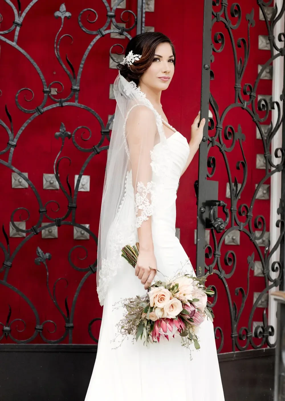 A bride in a white wedding dress holding a bouquet of flowers, standing by a decorative gate with a red background.
