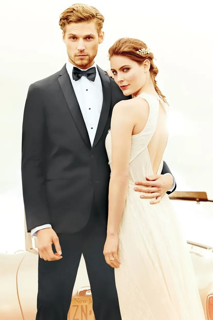 A couple in formal attire poses confidently. The man wears a black tuxedo with a bow tie, and the woman dons an elegant white dress, exuding romance and elegance against a light background.