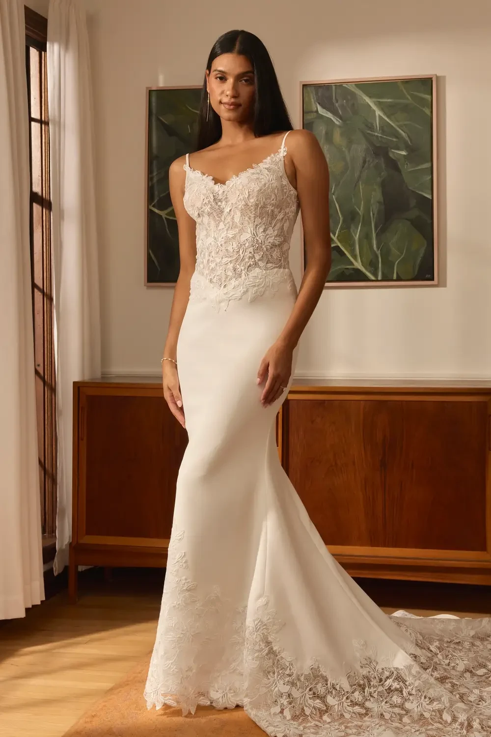 A woman in a fitted wedding dress with lace detailing standing in a softly lit room.