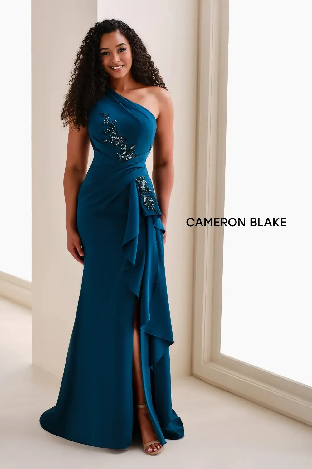A woman in a teal one-shoulder gown with decorative embellishments and a slit.