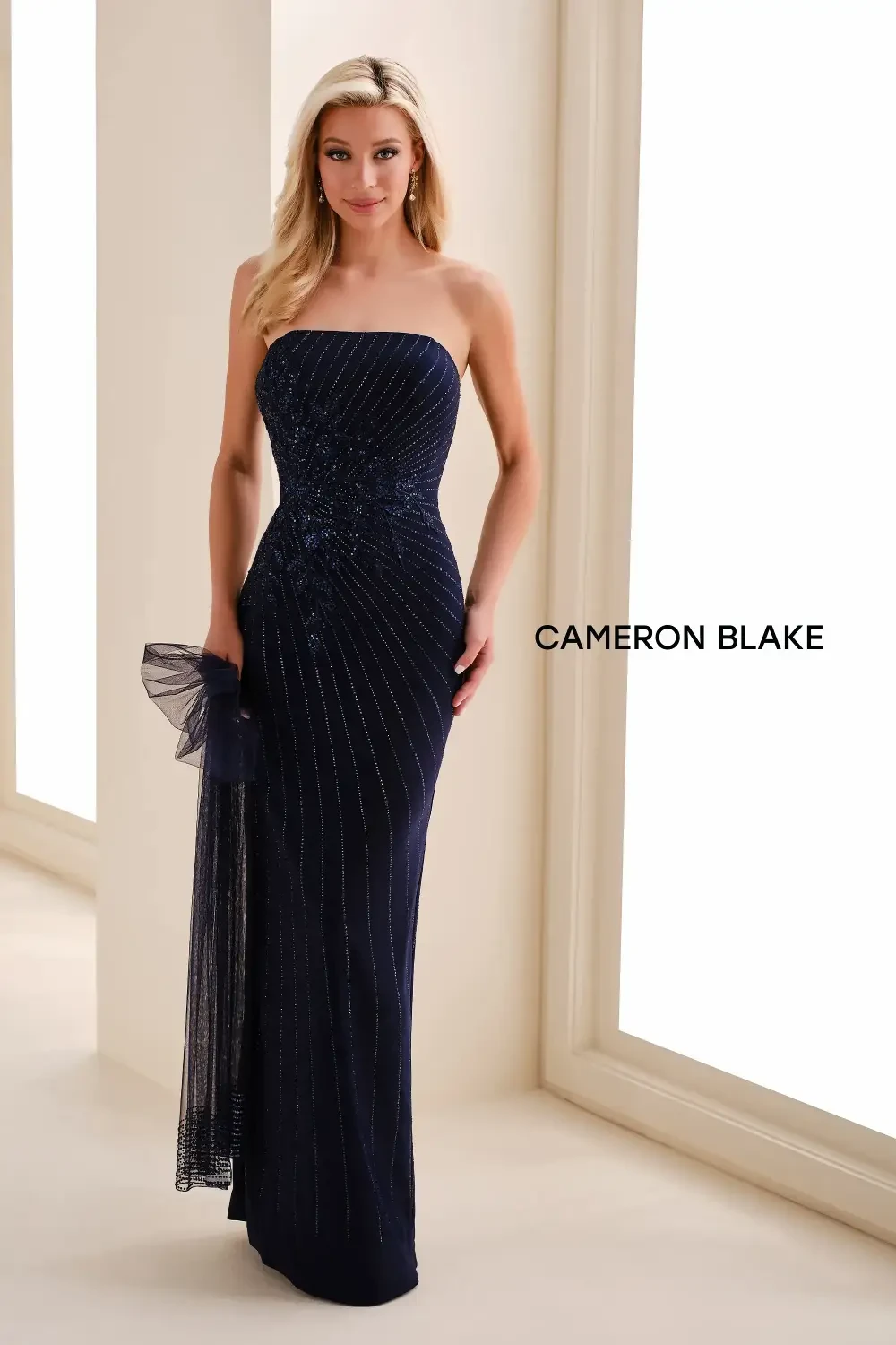 A woman in a strapless navy blue evening gown with a fitted silhouette and embellishments, standing in a well-lit space, accompanied by a sheer shawl.