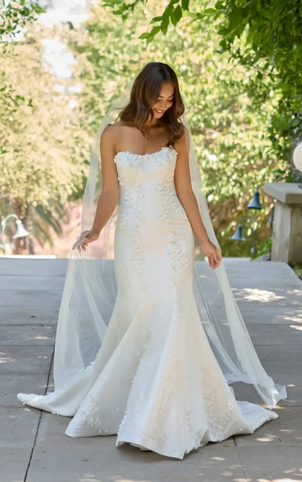 A woman in a white wedding dress with a fitted silhouette and a veil stands outside, surrounded by greenery.