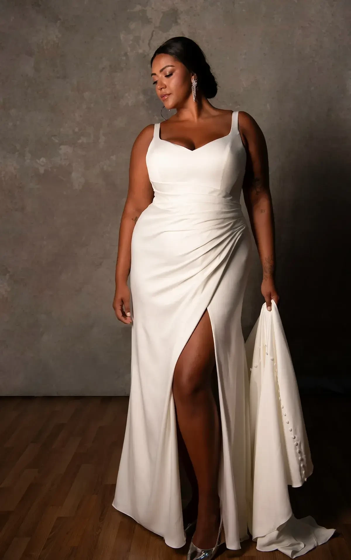 A woman in a white dress with a thigh-high slit, holding a fabric in one hand and posing confidently against a neutral background.