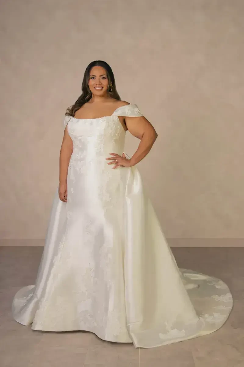 A woman in a white wedding dress with off-the-shoulder sleeves, standing and smiling. The dress has a long train and is set against a neutral background.