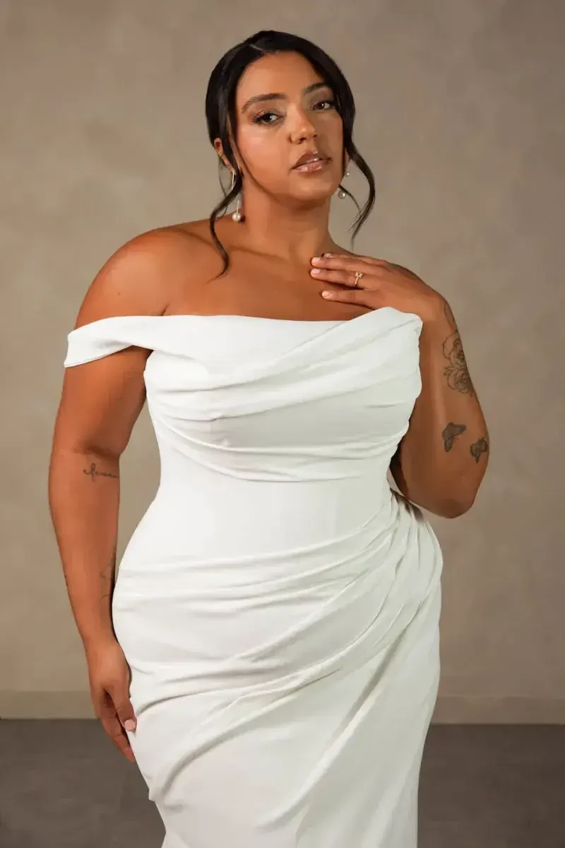 A woman posing in a form-fitting white off-the-shoulder dress with a textured fabric, standing against a neutral background. She has long dark hair and visible tattoos on her arms.