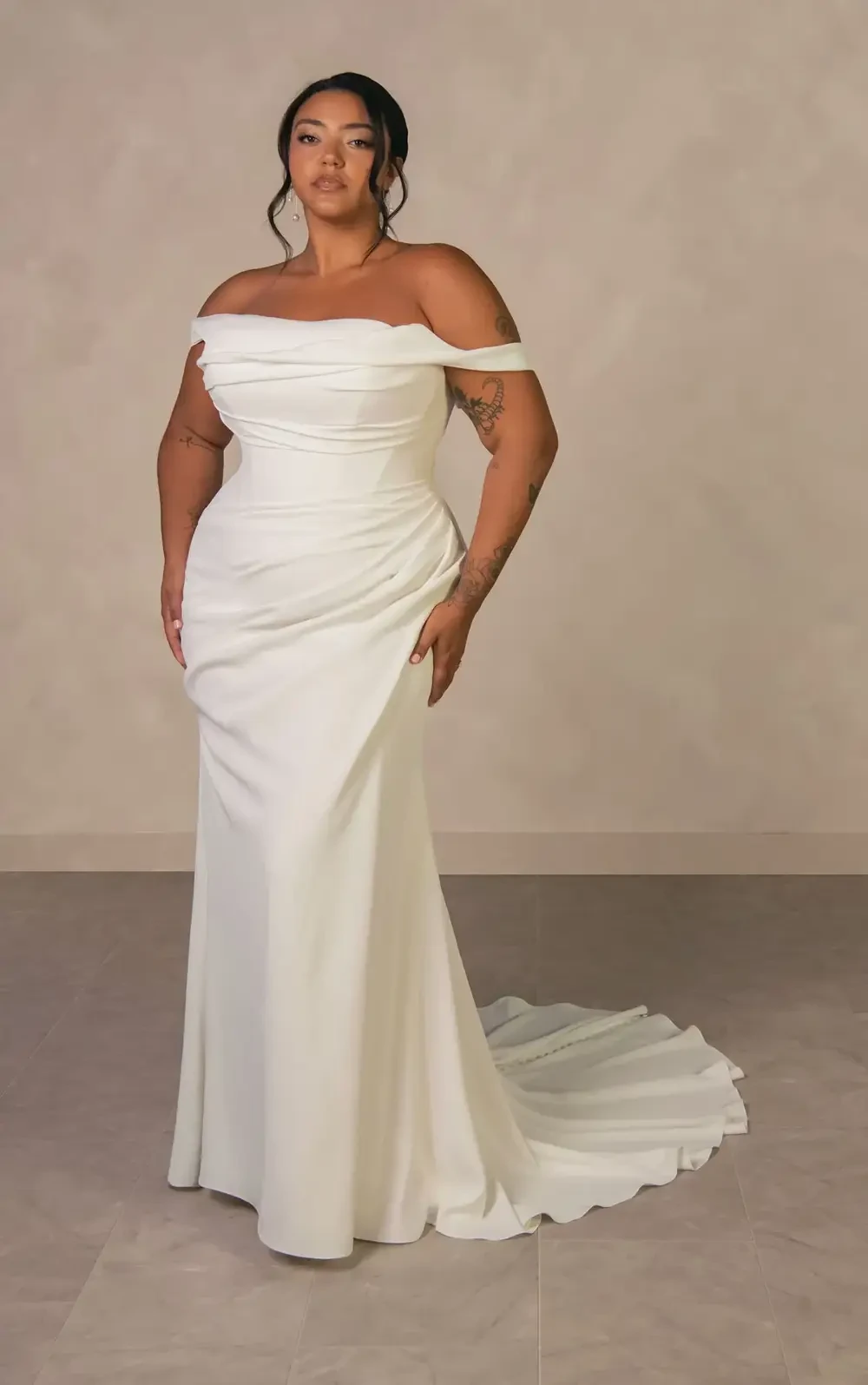 A woman wearing an elegant off-the-shoulder white gown, posing confidently against a neutral backdrop.