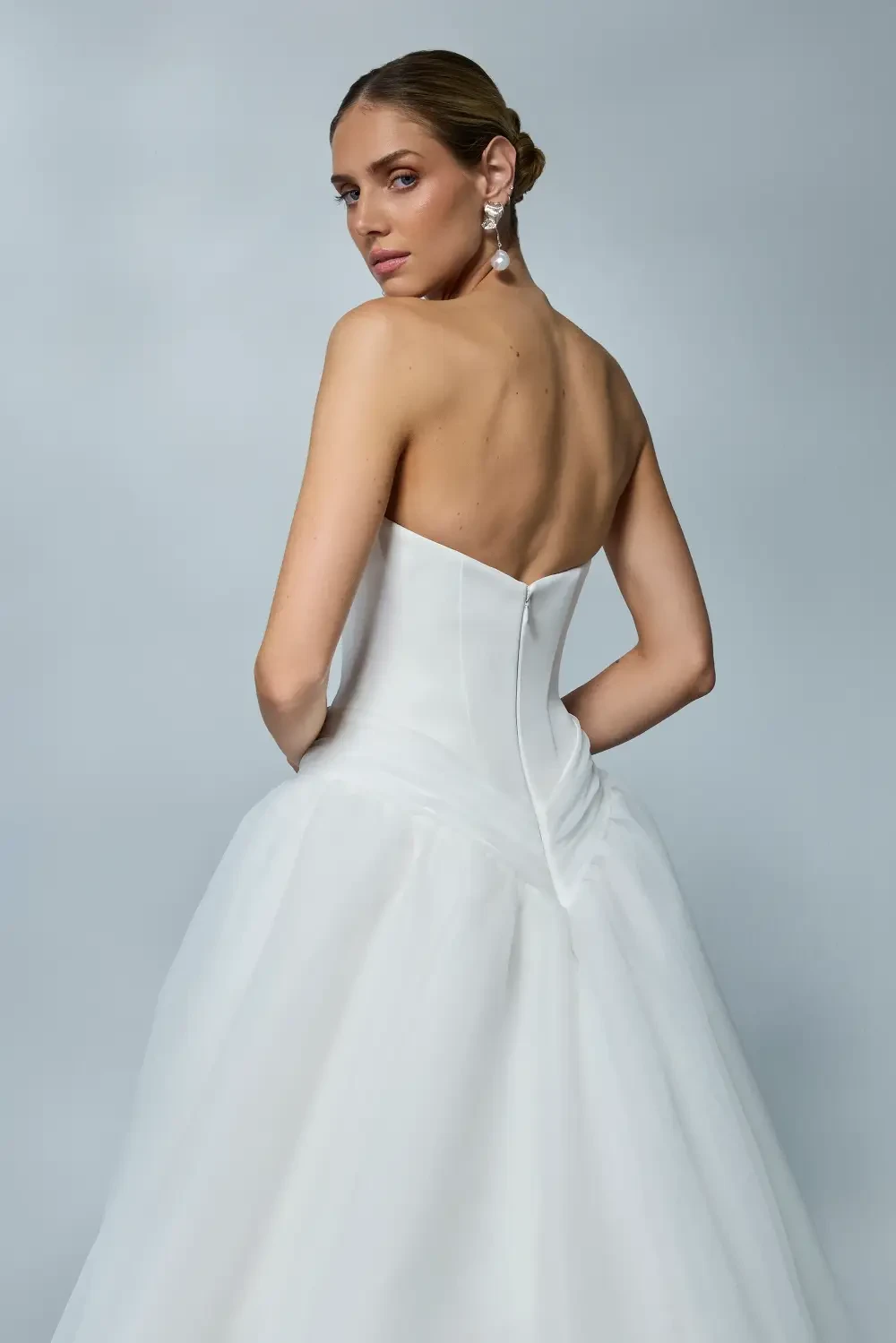 A woman wearing a strapless white wedding gown, posing from the back with an elegant hairstyle and earrings.