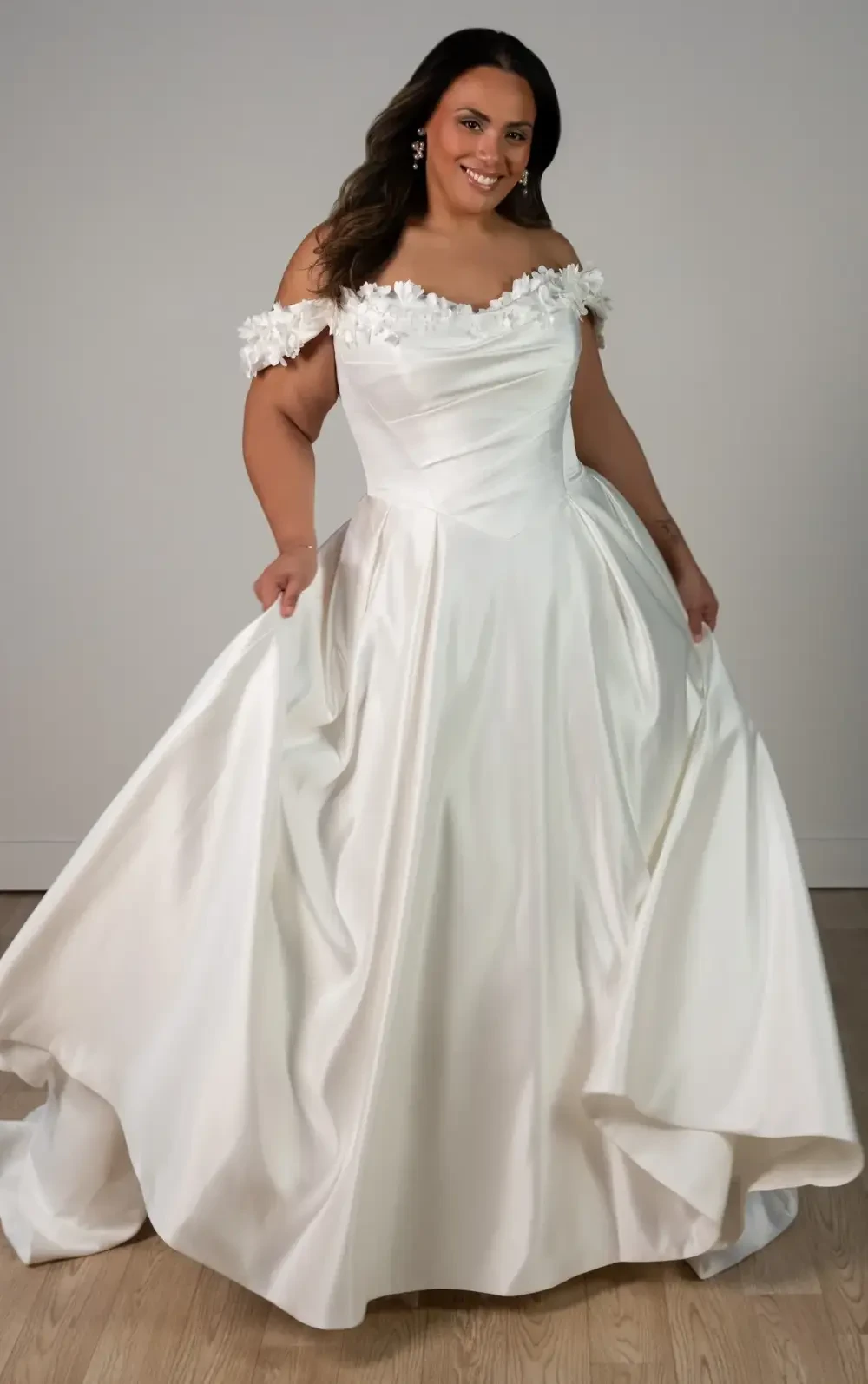 A person wearing a white wedding dress with off-the-shoulder sleeves and floral embellishments, smiling as they pose.