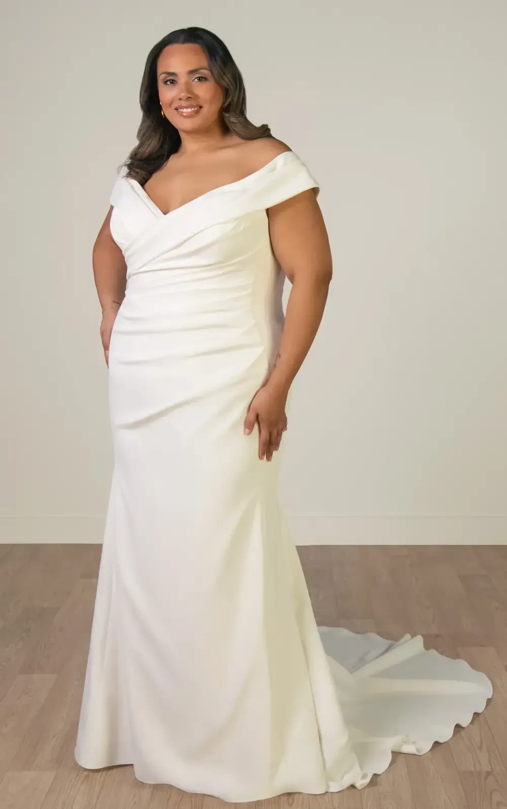 A woman in a white off-the-shoulder wedding gown stands confidently, smiling, against a neutral backdrop.